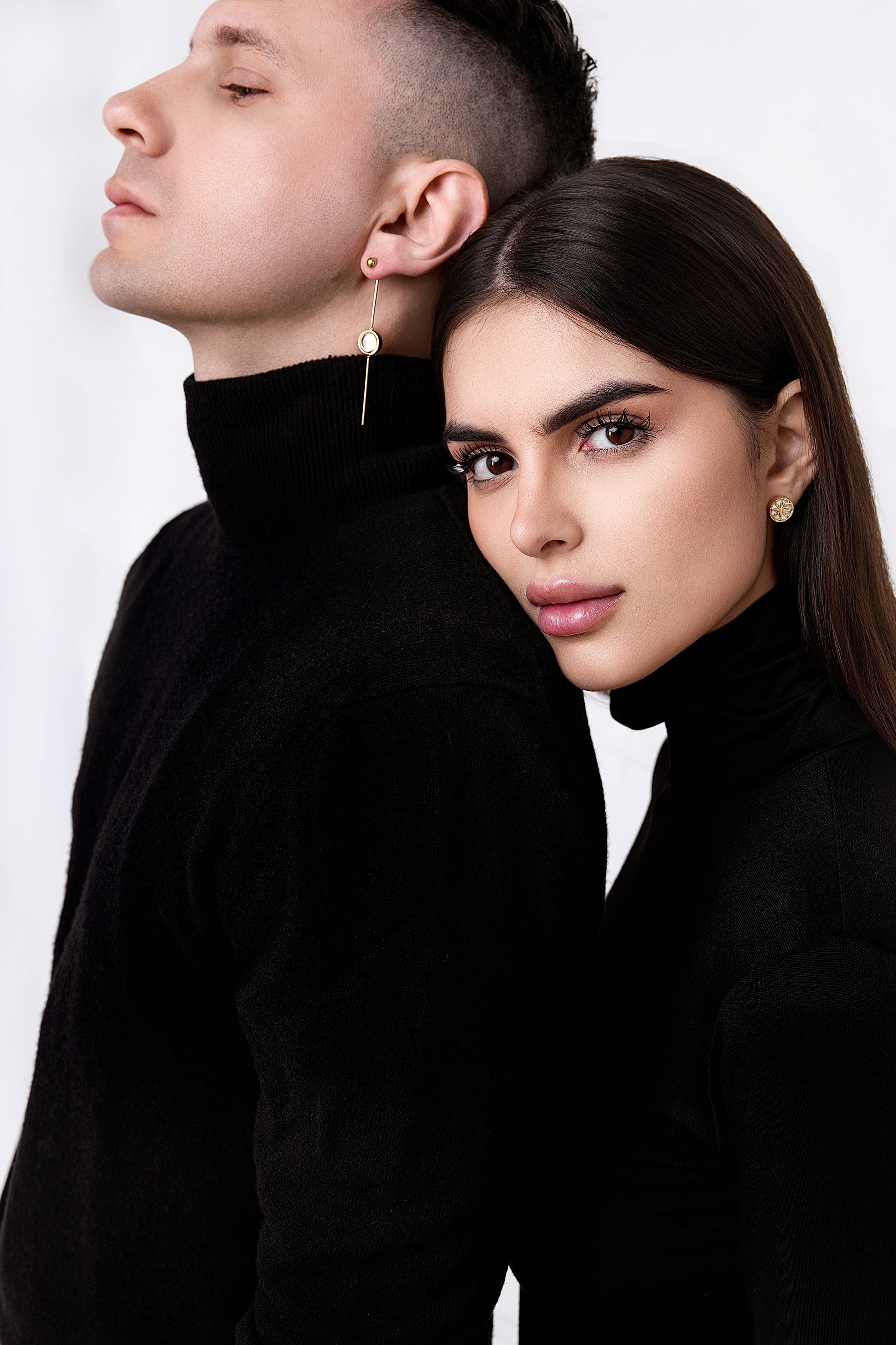 Man and woman standing close together with a plain background. She is wearing The Ravenna White Mother of Pearl Stud Earring in 18kt Yellow Gold. He is wearing the The Ravenna White Mother of Pearl 3-in-1 Long Earring in 18kt Yellow Gold.