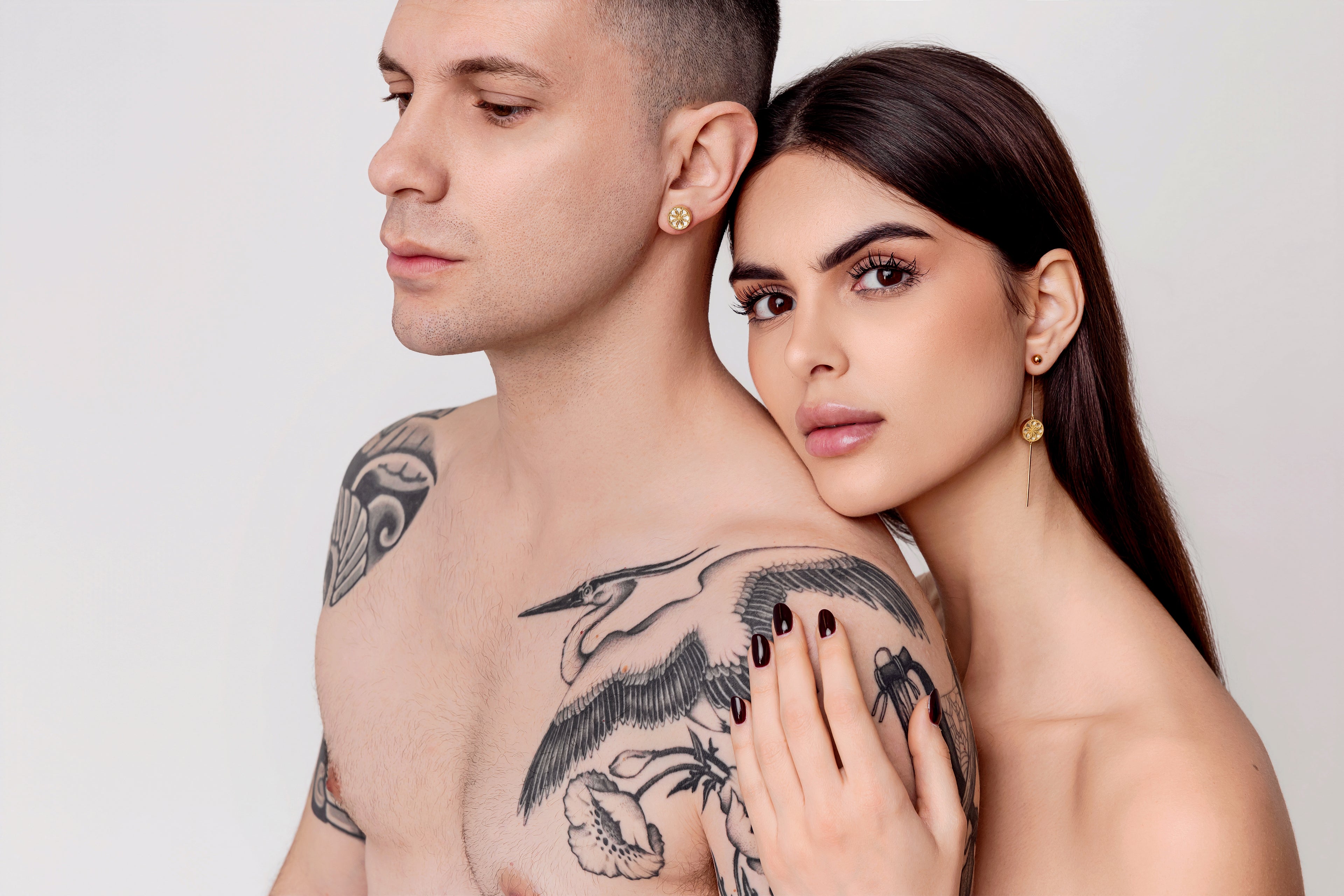 Man with tattoos on his shoulder and chest embracing a woman against a plain background. Intimacy and sharing jewels. The Ravenna White Mother of Pearl 3-in-1 Long Earrings in 18kt Yellow Gold. The Ravenna White Mother of Pearl Stud Earring in 18kt Yellow Gold.