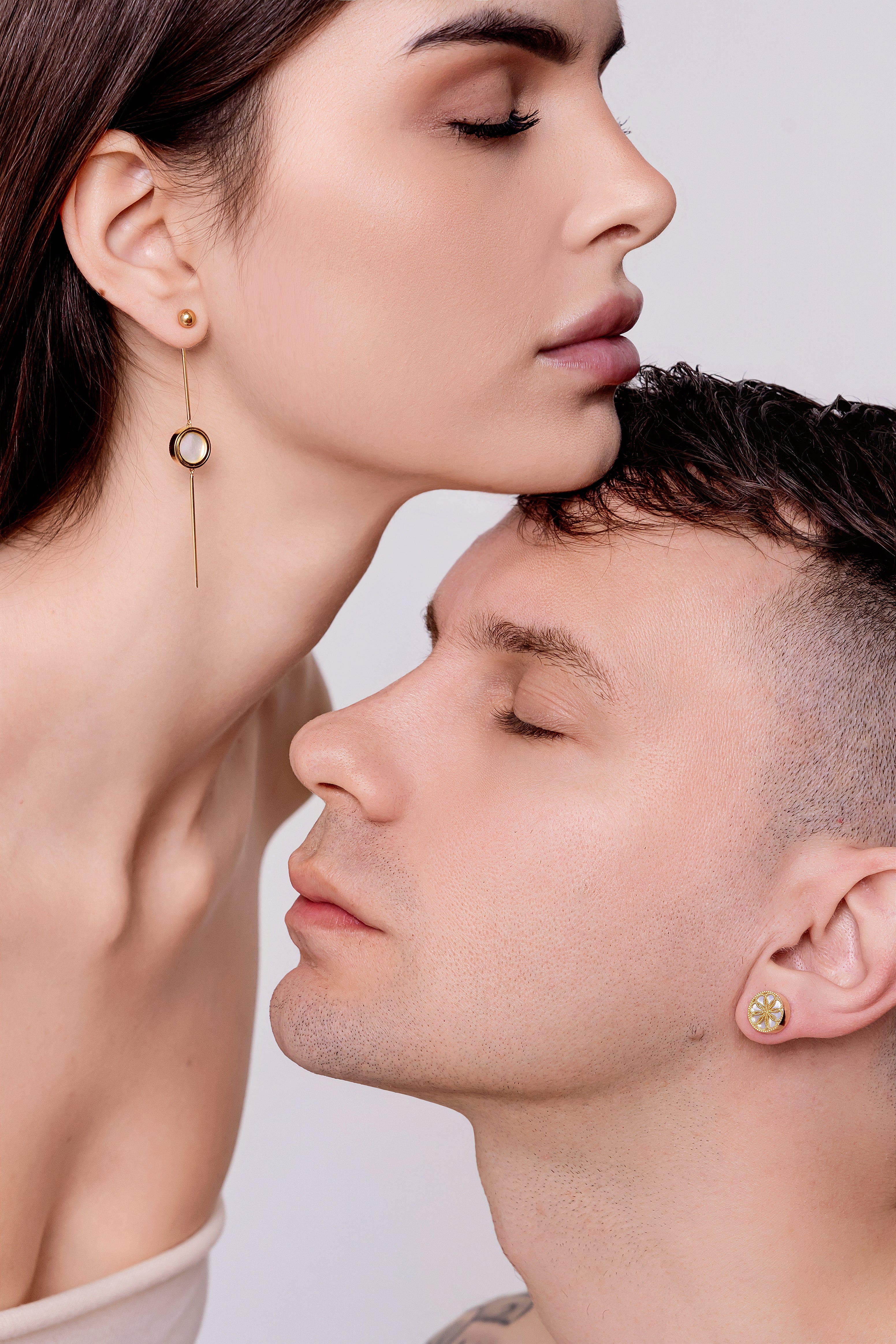 Close-up of a woman and a man wearing gold earrings against a neutral background. Two people wearing gold earrings against a white background. She is wearing The Ravenna White Mother of Pearl 3-in-1 Long Earrings in 18kt Yellow Gold. He is wearing the Ravenna White Mother of Pearl Stud Earrings in 18kt Yellow Gold. 