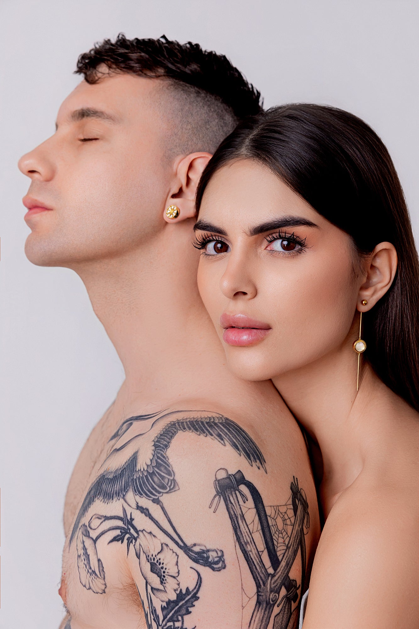 Man and woman standing close together with a plain background both wearing Earrings from Gió VELORI collection.