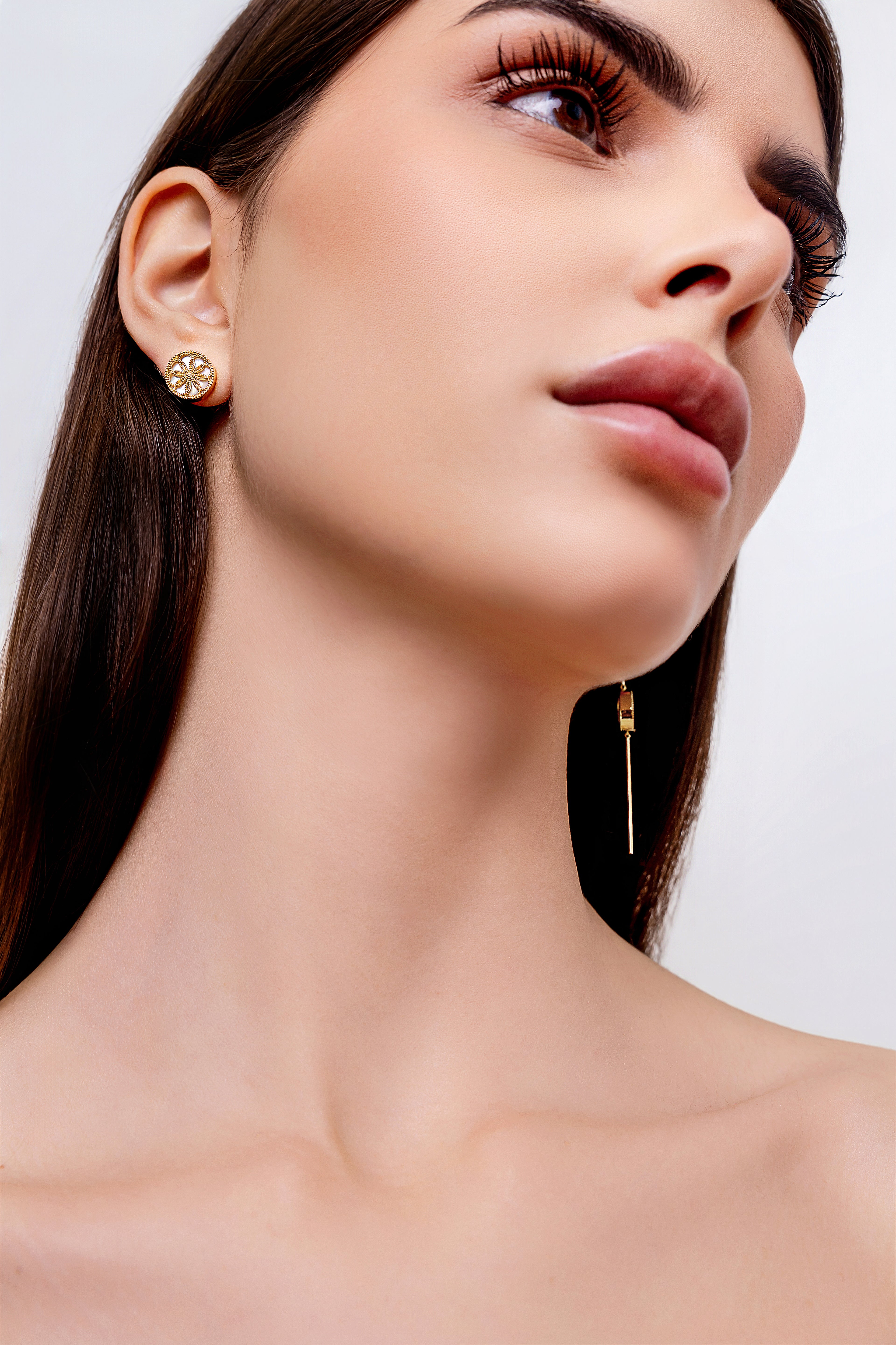 Close-up of a woman wearing gold earrings against a neutral background. The Ravenna White Mother of Pearl 3-in-1 Long Earrings in 18kt Yellow Gold and The Ravenna White Mother of Pearl Stud Earrings in 18kt Yellow Gold