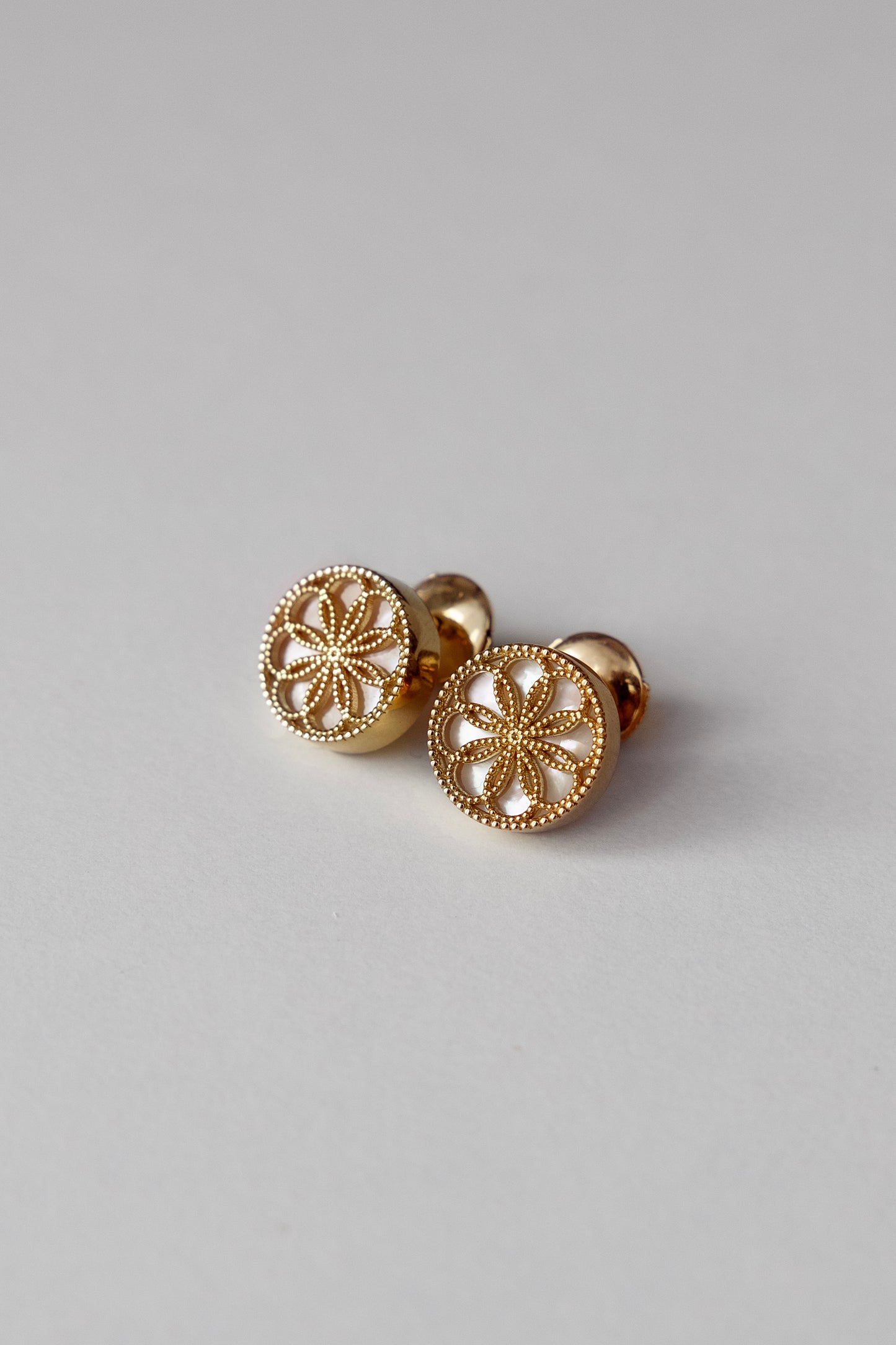 Gold earrings with a byzantine inspired design on a light grey background. The Ravenna White Mother of Pearl Stud Earrings in 18kt Yellow Gold.