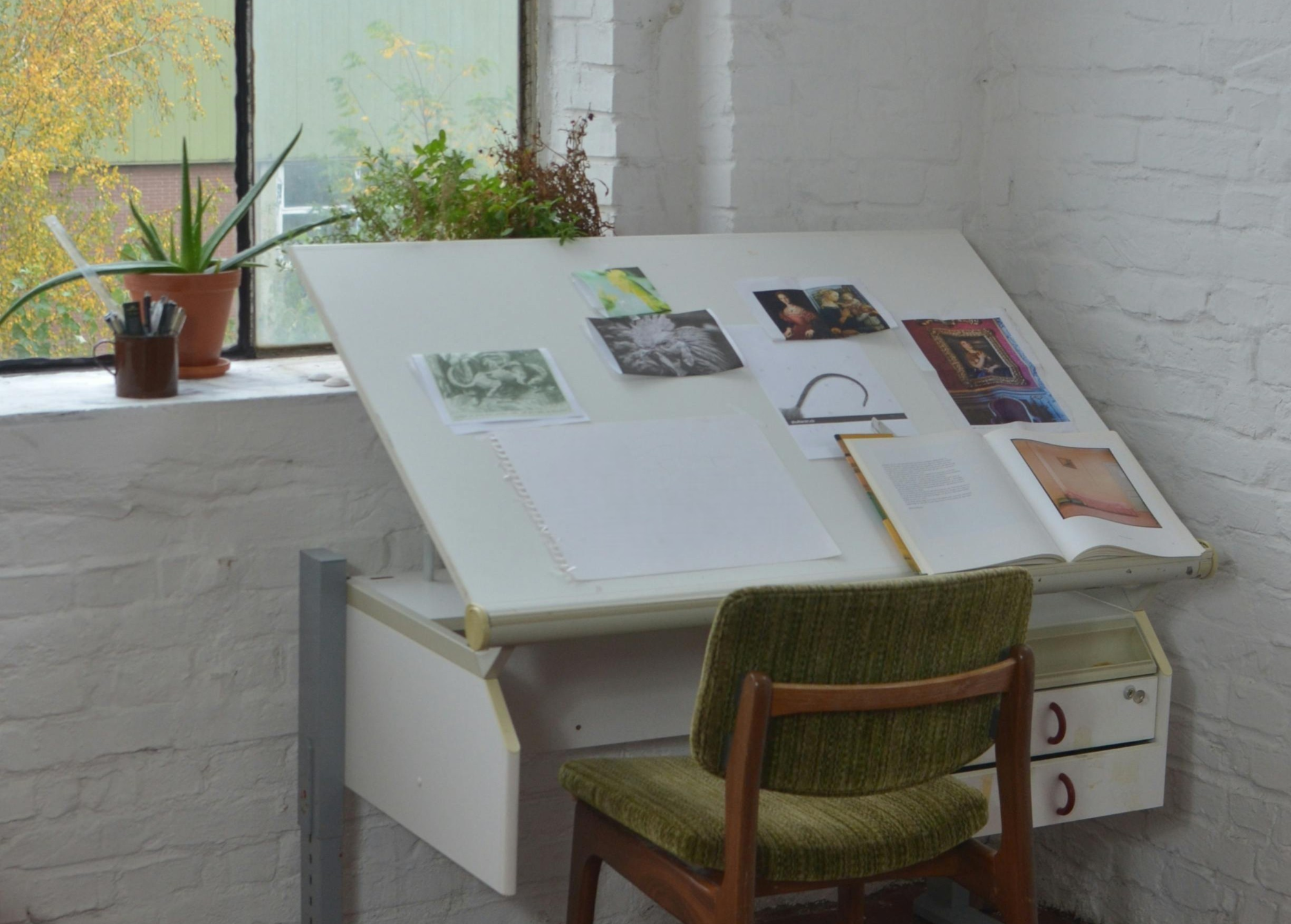 Artistic workspace with drawing table, chair, and plants in a room with white brick walls. Bespoke Orders. Made in italy.